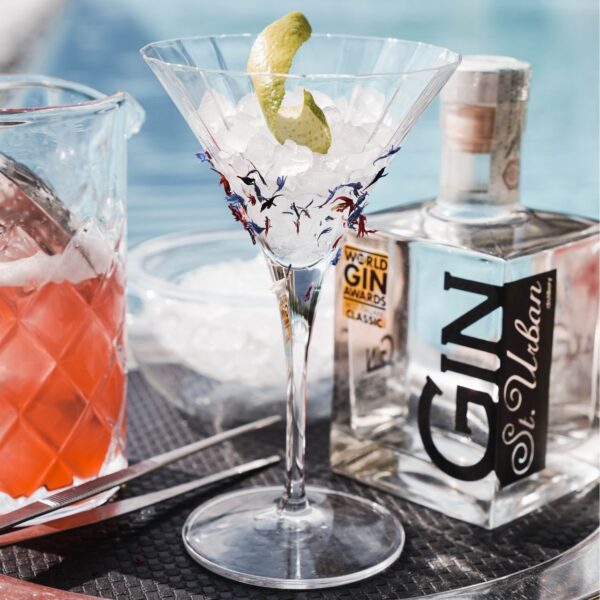 Martini glass filled with ice, citrus peel, and garnish next to a bottle of St. Urban Gin and a pink cocktail.