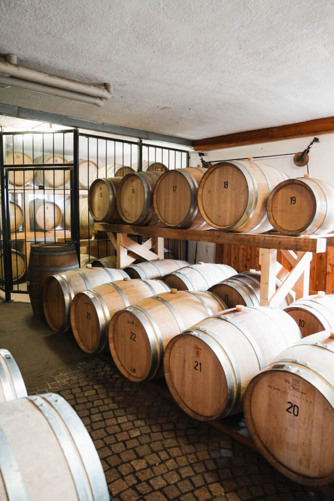 Wine cellar interior with rows of numbered oak barrels stacked on wooden shelves and a cobblestone floor.