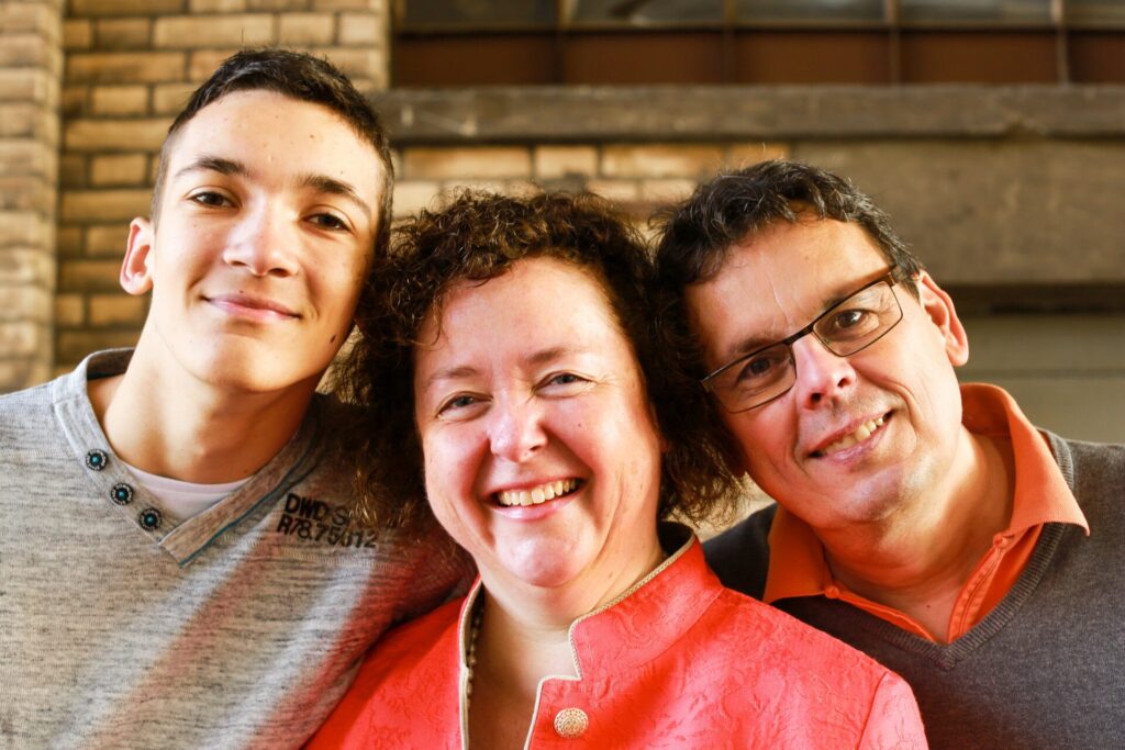 A smiling family of three poses closely together in front of a brick wall, all looking at the camera.
